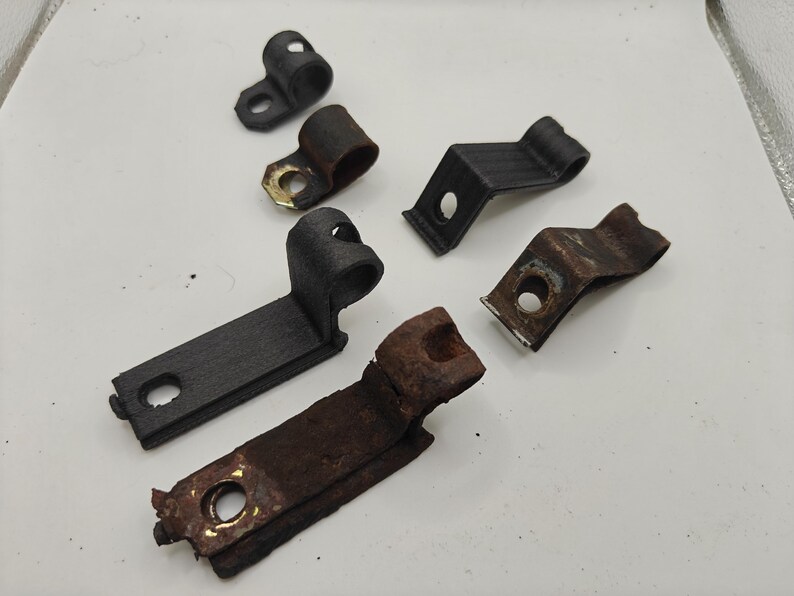 May include: A collection of six metal mounting brackets and clamps of varying sizes and conditions. Some pieces feature a smooth black finish, while others exhibit significant surface rust and oxidation, each with a single circular hole.