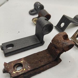 May include: A collection of L-shaped metal mounting brackets with a cylindrical loop at one end and a flat base with a circular hole. The set includes both matte black textured brackets and weathered, rusted brown metal brackets of identical shape.