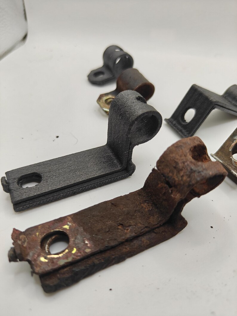 May include: A collection of L-shaped metal mounting brackets with a cylindrical loop at one end and a flat base with a circular hole. The set includes both matte black textured brackets and weathered, rusted brown metal brackets of identical shape.