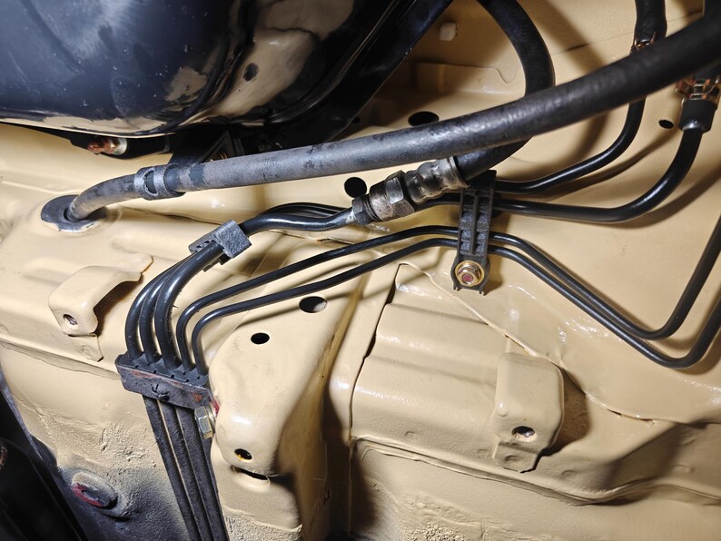 May include: A beige painted automotive undercarriage section featuring a series of thin black metal fuel or brake lines secured by black plastic mounting clips and a thick black rubber hose connected with a metal fitting to the chassis.