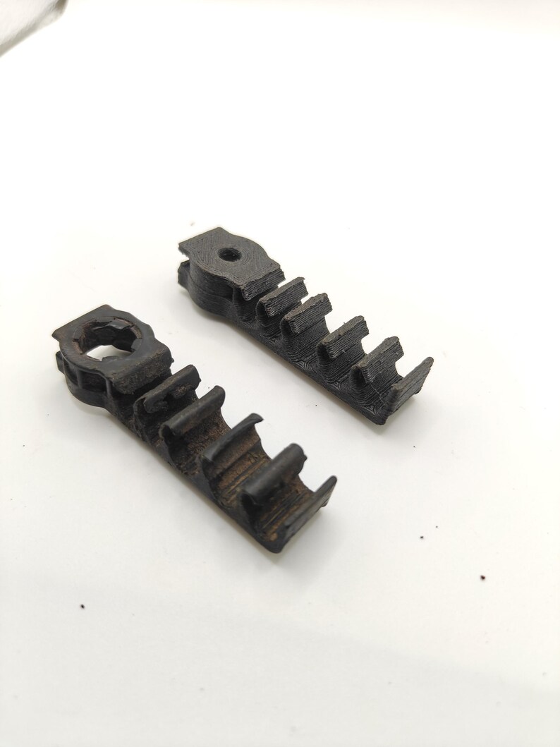 May include: Two black, 3D-printed plastic cable management clips. Each piece features a circular mounting hole at one end and a series of four open-ended, C-shaped channels designed to hold and organize individual wires or cables in a row.