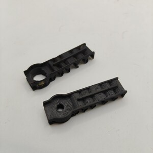 May include: Two black, 3D-printed plastic components with a rectangular body and a circular end. Each piece features a central hole and a series of small, evenly spaced vertical ridges along one side, showing a textured, layered surface finish.