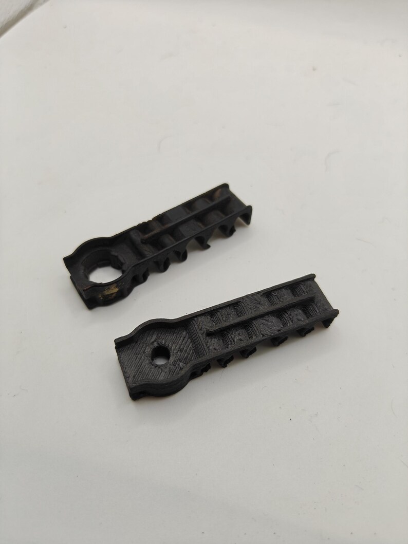 May include: Two black, 3D-printed plastic components with a rectangular body and a circular end. Each piece features a central hole and a series of small, evenly spaced vertical ridges along one side, showing a textured, layered surface finish.