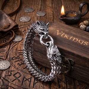 Può includere: Un bracciale a catena in metallo color argento con due terminali a forma di testa di lupo dettagliati che si collegano con una chiusura ad anello. Il bracciale presenta un design a maglie intrecciate spesse ed è appoggiato su una scatola di legno scuro con la scritta Vikings.