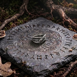 May include: A silver-toned ring shaped like a coiled snake with textured scales, resting on a square dark grey stone slab engraved with a circular Celtic knot pattern and a border of runic characters carved into the surface of the stone.