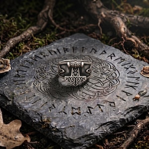 Op de afbeelding: Een zilverkleurige metalen ring met een gedetailleerd Mjolnir-hamerontwerp en ingewikkelde krulpatronen aan de voorzijde. De ring heeft een gepolijste afwerking met donkere, verdiepte delen die de gegraveerde details van de hamerkop benadrukken.
