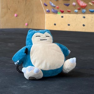 May include: A plush toy shaped like a round, blue and cream-coloured creature with closed eyes, a small mouth, and white claws on its hands and feet. The soft character is seated in a relaxed position with a black strap visible on its side.