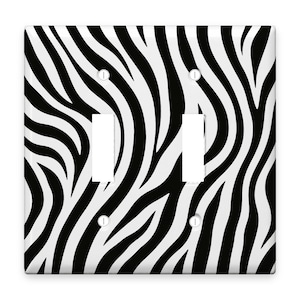 May include: A double light switch wall plate featuring a high-contrast black and white zebra stripe pattern. The rectangular cover includes two vertical rectangular cutouts for switches and four small circular holes for mounting screws.