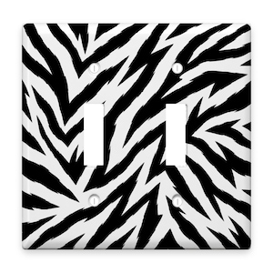 May include: A double light switch wall plate featuring a high-contrast black and white zebra stripe pattern. The rectangular cover includes two vertical rectangular cutouts for rocker switches and four small circular holes for mounting screws.
