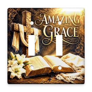 May include: A double light switch cover featuring a wooden cross draped in white fabric, an open Bible, and white lilies. The text Amazing Grace is printed in gold script against a dark, glowing background with ornate filigree details.
