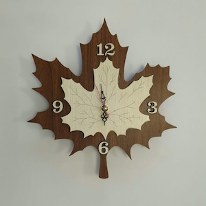 May include: A wall clock shaped like a maple leaf, featuring a dark brown outer layer and a lighter cream-colored inner layer with etched leaf veins. The clock face displays the numbers 12, 3, 6, and 9 in a serif font, with black ornate hands.