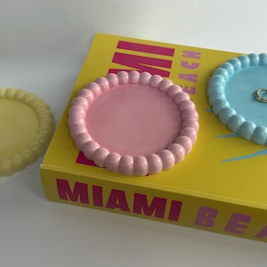 Può includere: Tre vassoi circolari per bigiotteria con bordi smerlati in giallo, rosa e azzurro. I vassoi rosa e azzurro sono posizionati su un libro giallo con il testo MIAMI BEACH, mentre il vassoio giallo si trova separatamente sulla sinistra.