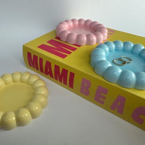Può includere: Tre vassoi circolari per gioielli con bordi smerlati a forma di bolla in giallo pastello, rosa chiaro e azzurro cielo. I vassoi rosa e blu poggiano su un libro giallo con il testo MIAMI BEACH, quello giallo è posizionato a parte.