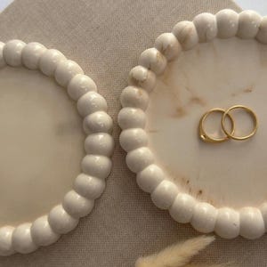 Può includere: Due vassoi circolari color bianco sporco con un bordo rialzato a perline e una superficie liscia e marmorizzata. Il vassoio di destra contiene due semplici anelli color oro. I vassoi presentano una consistenza arrotondata uniforme.