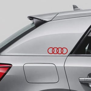 May include: A red Audi four-ring logo decal applied to the rear side window of a silver vehicle. The sticker features the iconic interlocking circular design in a solid red colour, positioned horizontally on the glass surface near the car body.