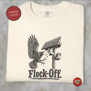 Puede incluir: Una camiseta color hueso con un gráfico en tinta negra de un cuervo volando hacia un poste de cámara de seguridad solar. El texto "Flock-Off." está impreso en una fuente negrita y desgastada debajo de la ilustración. Etiqueta Comfort Colors.