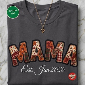 Puede incluir: Una camiseta gris oscuro con la palabra MAMA en letras grandes estilo patchwork con patrones otoñales que incluyen hojas, bellotas y cuadros. Debajo de la palabra, el texto dice Est. Jan 2026. Un collar con colgante de hoja dorado descansa encima.