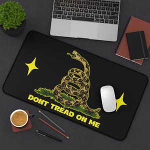 Könnte beinhalten: Eine rechteckige schwarze Schreibtischunterlage mit einer Grafik einer zusammengerollten Klapperschlange in Gelb und Braun sowie dem Text "DONT TREAD ON ME" in gelben Großbuchstaben. Zwei gelbe vierzackige Sterne flankieren das Motiv.
