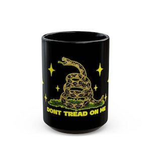 May include: A black ceramic mug featuring a coiled rattlesnake graphic in shades of brown and tan, positioned above green grass. The design includes yellow four-pointed stars and the yellow text DONT TREAD ON ME printed at the base.