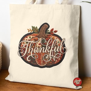 May include: A cream-colored canvas tote bag featuring a large, central embroidered pumpkin graphic. The pumpkin is filled with a patchwork of autumn-themed patterns in orange, red, and green, with the word Thankful written in white script.