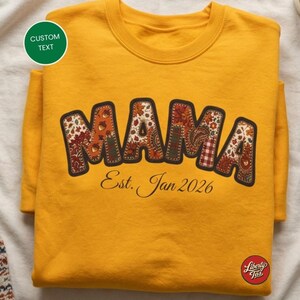 Peut inclure: Un sweat-shirt à col rond jaune moutarde avec le mot MAMA en grandes lettres majuscules remplies d'un motif floral et de glands aux couleurs automnales. Sous le mot, le texte Est. Jan 2026 est imprimé dans une police cursive noire.