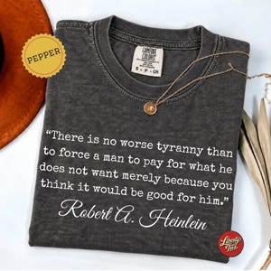 May include: A charcoal gray crew neck t-shirt featuring the white printed quote: "There is no worse tyranny than to force a man to pay for what he does not want merely because you think it would be good for him." by Robert A. Heinlein.
