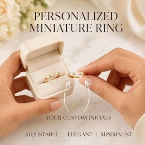 Puede incluir: Un anillo ajustable de tono dorado con dos extremos abiertos, cada uno grabado con la letra A o L. El anillo se presenta en una caja de color crema. El texto en la imagen dice: PERSONALIZED MINIATURE RING, YOUR CUSTOM INITIALS, ADJUSTABLE | ELEGANT | MINIMALIST.