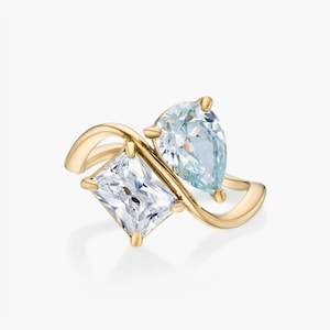 Pode incluir: Um anel de ouro com duas pedras proeminentes em estilo bypass. Uma pedra é uma gema transparente de corte radiante retangular e a outra é uma gema azul clara em forma de pera. A banda de ouro polido curva-se graciosamente em torno delas.