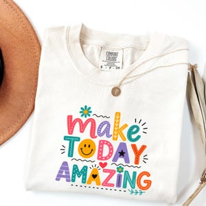Puede incluir: Una camiseta de color crema con el texto colorido "make TODAY AMAZING", una cara sonriente amarilla, un pequeño corazón rojo y detalles florales. La camiseta tiene una etiqueta de la marca Comfort Colors en el cuello y un collar dorado fino.