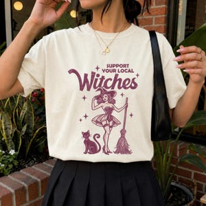 May include: A cream-colored short-sleeve t-shirt featuring a dark purple graphic print. The text reads "SUPPORT YOUR LOCAL Witches" above an illustration of a person in a witch hat and corset holding a broom, accompanied by a black cat and stars.