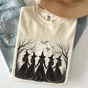 May include: A cream-colored t-shirt featuring a black ink illustration of five witches in pointed hats holding hands in a circle under a large full moon, surrounded by bare trees. The inner neck label displays the text Comfort Colors.