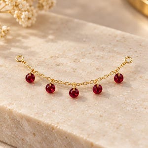 May include: A gold-toned chain connector featuring five small, round, faceted red crystals dangling from individual jump rings. The chain has a circular loop at each end for attachment, creating a delicate, symmetrical jewelry component.