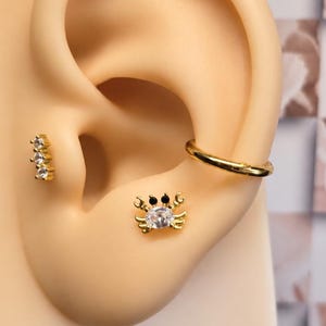 May include: A gold-toned ear jewelry set featuring a crab-shaped stud with a clear center stone and black eyes, a three-stone vertical bar stud with clear crystals, and a plain gold-toned hoop earring, all displayed on a neutral ear model.