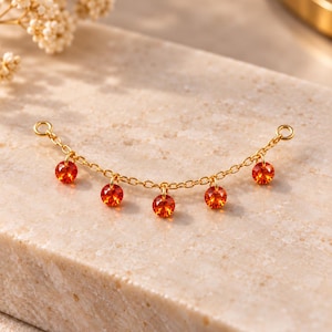 May include: A gold-toned chain connector featuring five small, round, faceted orange crystals suspended from individual jump rings. The delicate metal chain has a circular loop at each end, designed for attachment to other jewelry components.