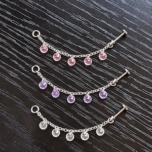 May include: Three silver-toned body jewelry chains, each featuring a flat-back post on one end and a circular ring on the other. The chains are adorned with five dangling round-cut gemstones in shades of pink, purple, and clear white.