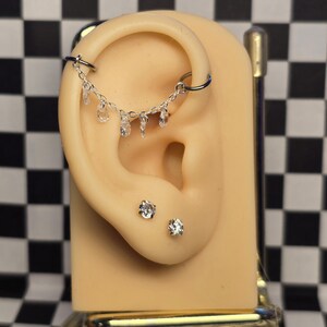 May include: A silver-toned ear cartilage hoop connected by a delicate chain featuring five small, clear, teardrop-shaped dangling crystals. Below the hoop, two separate round, clear stud earrings are positioned on the earlobe.