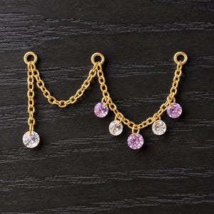 May include: A gold-toned metal chain connector featuring three circular jump rings at the top. The chain drapes in two sections, adorned with six dangling round-cut gemstones in alternating clear and light purple colors.