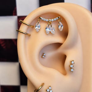 May include: A gold-toned ear curation featuring a curved barbell with three marquise-cut clear stones, two plain gold hoops, a three-stone cluster stud, a vertical three-stone bar stud, and a single gold hoop with a three-stone cluster stud.