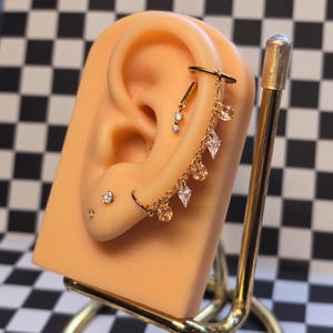 May include: A gold-toned ear cuff connected by a delicate chain to a lower hoop earring, featuring dangling round and diamond-shaped crystals. Additional small stud earrings are placed in the earlobe and cartilage area.