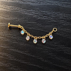 May include: A gold-toned body jewelry piece featuring a flat-back labret stud connected to a delicate chain. The chain is adorned with five small, round, iridescent crystals that dangle from the links, creating a shimmering, multi-faceted effect.