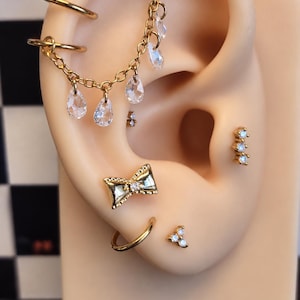 May include: A gold-toned ear display featuring multiple piercings, including two plain hoops, a chain with dangling clear teardrop crystals, a bow-shaped stud with a central stone, a three-stone cluster stud, and a small triangular stone cluster.
