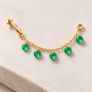 May include: A gold-tone body jewelry piece featuring a flat-back post connected to a delicate chain. The chain is adorned with five small, round, faceted green stones that hang as charms, creating a curved, dangling design.