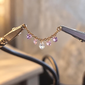 May include: A gold-toned chain segment featuring five dangling faceted gemstones. The center stone is a clear, pear-shaped gem, flanked by two smaller round pink stones on each side, all suspended from a delicate metal link chain.