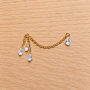 May include: A gold-toned metal chain connector featuring four dangling clear, teardrop-shaped crystals. The design consists of a central chain segment connecting two jump rings, with three crystals suspended from one side and one crystal on the other.