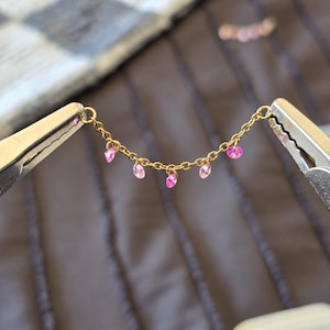 May include: A gold-toned metal chain featuring five small, faceted pink crystals suspended at regular intervals. The delicate chain is held taut by two metal clamps, showcasing the bright pink stones against the metallic gold links.