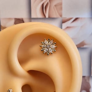 May include: A gold-toned ear piercing jewelry set featuring a central flower-shaped stud with marquise-cut clear stones, a smaller vertical bar stud with four clear stones, and a plain gold-toned hoop ring, all displayed on an ear model.