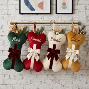 May include: Four bone-shaped Christmas stockings in dark green, red, cream, and gold velvet. Each stocking features a central ribbon, a large bow, and a personalized name embroidered in script: Ava, Emma, Noah, and Liam. They hang from a gold rod.