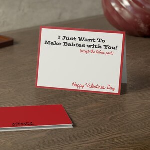 May include: A Valentine's Day card with the text "I Just Want To Make Babies with You! (except the babies part)" in black and red. A second red card is also visible. The cards are on a wooden surface.