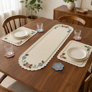 Pode incluir: Um caminho de mesa de crochê na cor creme e dois jogos americanos retangulares combinando, cada um com bordas festonadas e motivos florais bordados em rosa, azul e verde. O conjunto inclui quatro porta-copos de crochê em formato de flor.