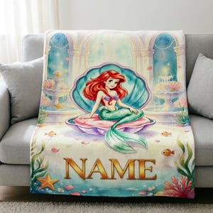 May include: A soft throw blanket featuring a watercolour illustration of a mermaid with long red hair sitting inside a large open seashell. The background shows an underwater palace with fountains, starfish, and coral. The word NAME is printed below.